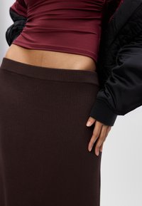 Kobieta w bordowej bluzce i brązowej, prążkowanej spódnicy, z widocznym rękawem czarnej kurtki, trzymająca rękę na biodrze.