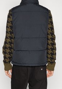 Marinblå puffväst med hög krage, med horisontell sömnad och en slät textur, lagd över en mönstrad stickad tröja i olivgrönt och svart.