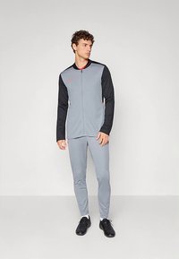 Pelēks sporta tērps ar melnām piedurknēm, ietver zip-jaku un piegulošas bikses. Apģērbu papildina melnas sporta kurpes.