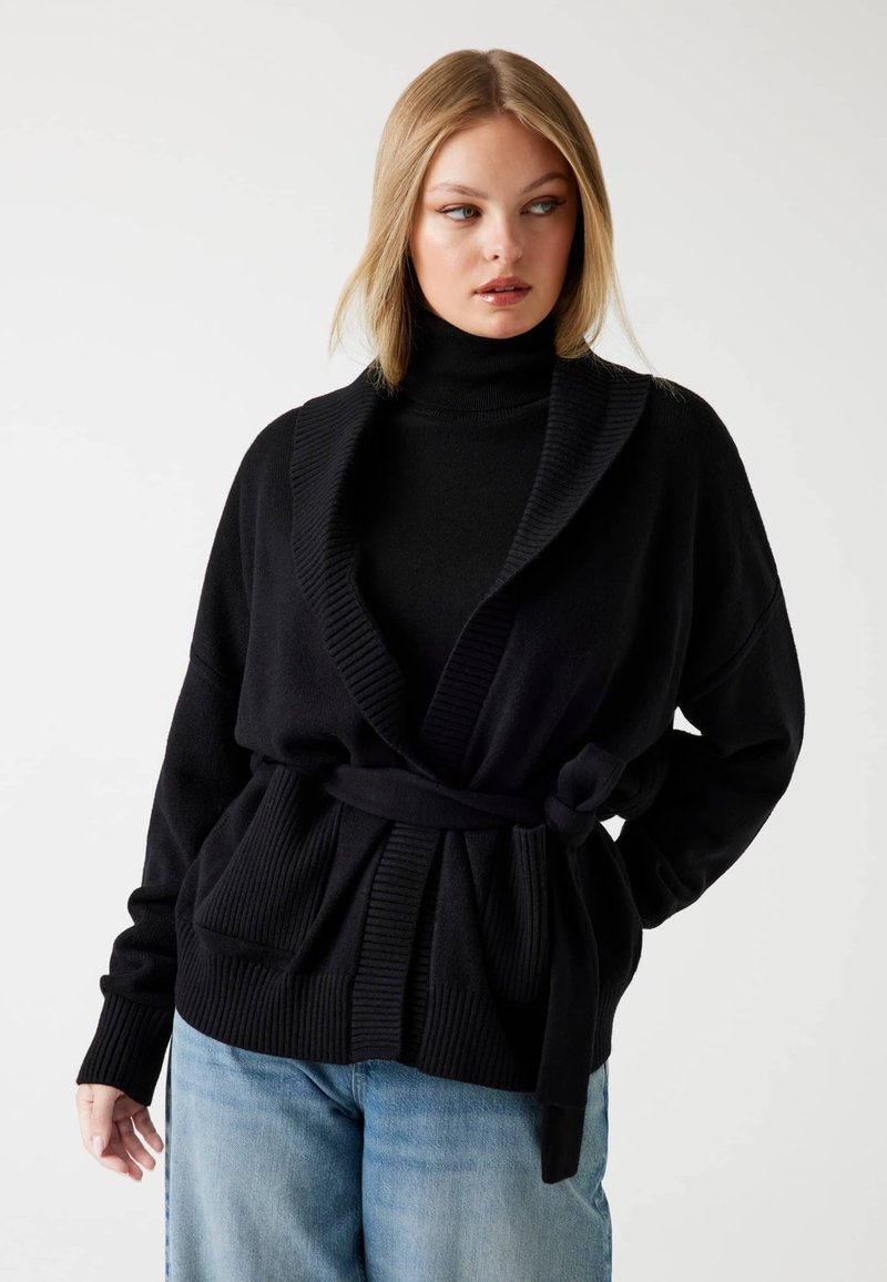 Sort cardigan med sjalskrave, bælte i taljen og ribbet tekstur. Har overdimensionerede lommer og lange ærmer. Varm, strikket stof.
