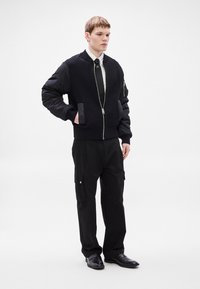 Casaco bomber preto com textura canelada, fecho de correr e bolsos; combinado com calças cargo largas pretas e sapatos pretos.