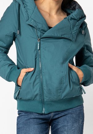 Personne portant une veste zippée couleur sarcelle avec capuche et poignets côtelés, mains dans les poches, associée à un jean bleu.