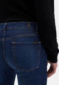 Ciemnoniebieskie jeansy z denimu o prostym kroju, pomarańczowym przeszyciu oraz metkami marki na pasku i tylnej kieszeni. Gładka faktura.