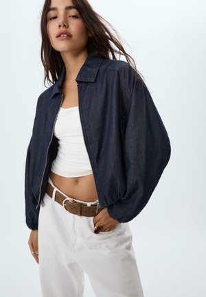 Frau trägt eine dunkle Jeansjacke mit Reißverschluss, ein weißes Crop-Top, weiße Hosen mit braunem Gürtel, eine Hand in der Tasche, stehend vor einem schlichten hellen Hintergrund.