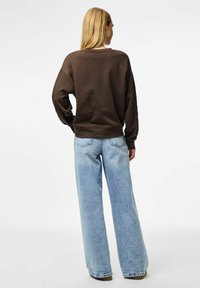 Sudadera marrón con puños y dobladillo acanalados, combinada con unos vaqueros de pierna ancha en azul claro. La tela parece suave con un corte informal.