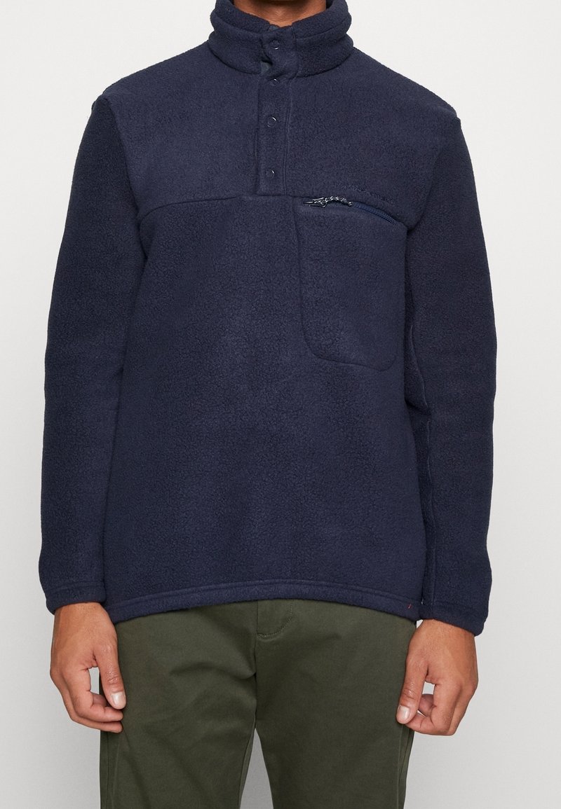 Sudadera polar azul marino con cuello alto, cierre de botones y un bolsillo en el pecho con cremallera. Tela texturizada con un aspecto suave.