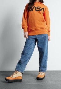 Orange hoodie with black NASA logo, loose blue denim jeans, and tan ankle boots with black rubber soles and lacing detail.