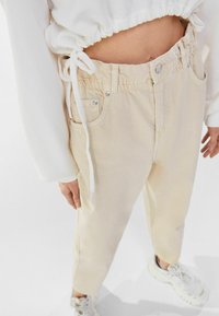 Kvinna som bär beige högmidjade avslappnade byxor, en kort vit huvtröja och vita sneakers, stående framför en vit bakgrund.