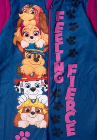 Tuta in pile con quattro cuccioli di cartone impilati, principalmente blu con dettagli rosa; la scritta recita "Feeling Fierce" (Sentirsi Feroce), con motivi di impronte di zampa.