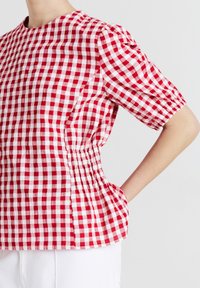 Blusa a maniche puff corte in gingham rosso e bianco con dettaglio in vita elastico, indossata con pantaloni bianchi su uno sfondo semplice.