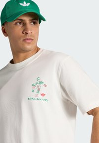Uomo che indossa una maglietta bianca con grafica di un giocatore di calcio in verde e rosso e testo "ITALIA '90", e un cappellino verde Adidas con logo bianco.