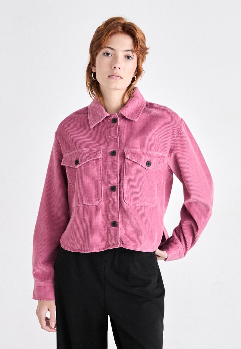 Růžová kordová bunda s cropped designem, předním zapínáním na knoflíky a dvěma náprsními kapsami. Obsahuje límec a dlouhé rukávy.