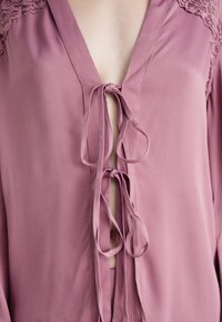Dettaglio ravvicinato di una blusa malva con spalle in pizzo e due fiocchi legati lungo il collo.