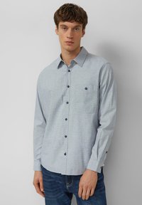 Lichtblauw overhemd met lange mouwen, een button-down kraag, een borstzak en donkere knopen; gemaakt van een gestructureerde stof. Draagt donkere spijkerbroek.