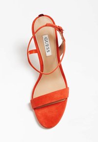 Rød ruskinds sandal med åben tå, ankelslynge og spænde, beige indersål mærket GUESS, fotograferet på en hvid baggrund.