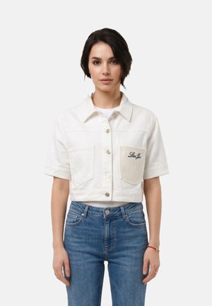 Donna che indossa una camicia bianca corta con bottoni, con una tasca destra texturizzata e jeans blu, in piedi davanti a uno sfondo chiaro e uniforme.