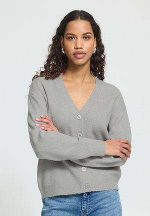 Vrouw met krullend haar, zilveren oorbellen, een grijze, dichtgeknoopte cardigan en blauwe jeans, met gekruiste armen staand tegen een effen achtergrond.