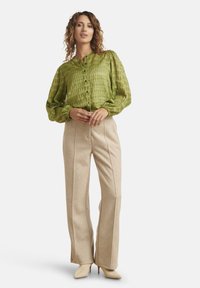 Blusa verde strutturata con bottoni frontali e maniche arricciate, abbinata a pantaloni beige su misura e tacchi chiari a punta.