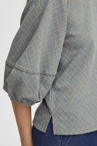 Blouse bleu clair à motifs avec des manches bouffantes. Présente un design géométrique en beige, un tissu texturé et des fentes sur les côtés pour la ventilation.