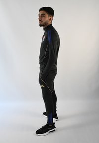 Chándal deportivo en negro con rayas azules y detalles dorados, con cuello alto y ajuste entallado. Zapatillas negras completan el look.