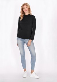 Fekete, hosszú ujjú, garbós felső világoskék, testhezálló farmerrel és fehér tornacipővel egy modellen, sima háttér előtt.