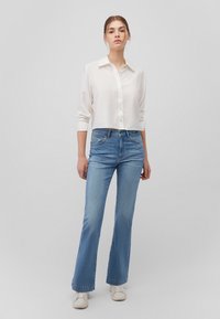 White, button-up, cropped shirt with long sleeves paired with light blue flared jeans and white sneakers, set against a neutral background.