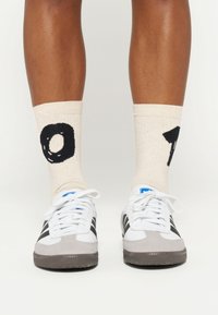 Białe sneakersy z szarymi zamszowymi akcentami, czarnymi paskami i niebieskim logo; noszone z kremowymi skarpetkami o prążkowanej fakturze, z dużymi czarnymi cyframi.