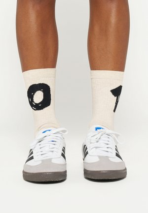 Białe sneakersy z szarymi zamszowymi akcentami, czarnymi paskami i niebieskim logo; noszone z kremowymi skarpetkami o prążkowanej fakturze, z dużymi czarnymi cyframi.