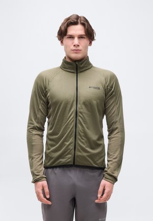 Giovane uomo con giacca verde oliva con cerniera e pantaloni grigi, in piedi davanti a uno sfondo semplice di colore grigio chiaro.