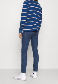 Camicia a maniche lunghe blu a righe con strisce arancioni, abbinata a jeans scuri aderenti e sneaker bianche alte. Vista da dietro.