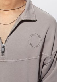 Světlě šedá mikina na zip vyrobená z měkké látky, vybavená čtvrtinovým zipem a kruhovým logem "Young Poets" na hrudi.