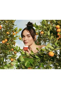 Un pot de crème à l'orange tenu parmi des feuilles vertes et des oranges ; une personne aux longs cheveux sombres regardant à travers le feuillage, montrant une peau lisse et une tenue neutre.