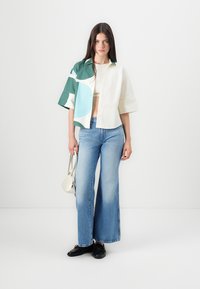 Camicia a maniche corte color crema con motivi astratti verde e blu, abbinata a jeans flare a vita alta di un blu chiaro e scarpe nere.