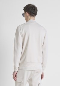Sudadera beige claro con cuello, que presenta una textura suave y puños acanalados. La parte posterior muestra un diseño limpio sin logotipos ni patrones visibles.