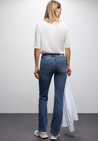 Jeans in denim blu con una vestibilità dritta, caratterizzati da una vita a media altezza, due tasche posteriori e un'etichetta in vita. Indossati con una maglietta bianca.