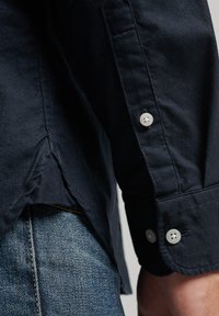 Navy button-up shirt featuring a smooth texture, two buttons on the cuff, and a curved hem. Paired with light blue denim jeans.