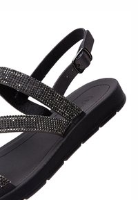 Sandale noire avec deux bandes croisées ornées de petits strass. Se règle avec une boucle latérale. Semelle intérieure texturée pour le confort.