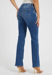 Jean en denim bleu foncé avec une coupe slim, des jambes droites et une taille basse. Présente un logo en triangle sur la poche arrière et un léger effet usé.
