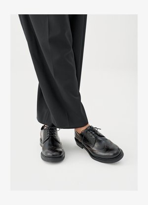 Schwarze Lederschuhe mit spitzer Zehe, dekorativen Perforationen und braunen Nähten. Kombiniert mit lockeren schwarzen Hosen.