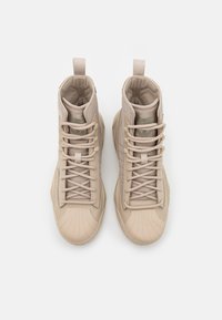 Sneaker alte beige con una toma superiore in tessuto testurizzato, accenti metallici, sistema di allacciatura prominente e suola in gomma con design curvo.