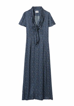 Femme portant une veste en cuir marron par-dessus une robe midi bleue à motifs, des chaussettes grises, des baskets bordeaux et un sac à bandoulière beige, debout sur un fond uni.