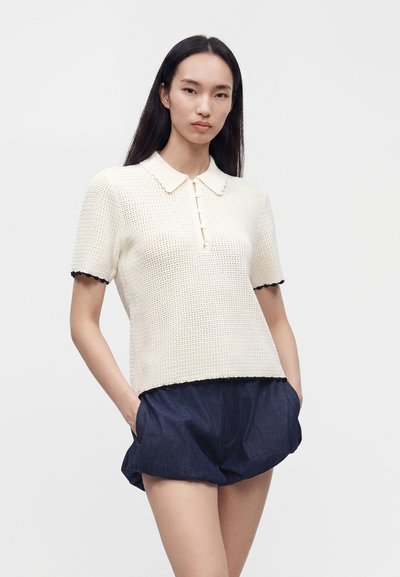 Femme aux longs cheveux noirs portant un polo en maille crème à manches courtes et un short bleu foncé ample, debout les mains dans les poches devant un fond uni.