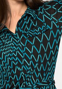 Svart skjorta med en teal-färgad diamantmönsterdesign. Har en krage och en ficka på vänster sida. Strukturerad tyg.