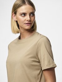 Žena s kratkom plavom kosom, noseći bež majicu s okruglim izrezom, gleda lijevo na jednobojnoj bijeloj pozadini.