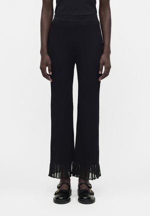 Άτομο που φοράει μαύρο ριμπ πλεκτό παντελόνι με πιετέ κρόσσια στο τελείωμα και μαύρα παπούτσια Mary Jane με χρυσά αγκράφες, στέκεται μπροστά σε λευκό φόντο.