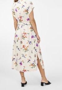 Floral midi-jurk van crèmekleurige stof, met paarse en oranje bloemen, korte mouwen, zijsplitten en een getailleerde taille. Zwarte blokhakken sandalen.