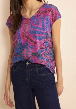 Vrouw draagt een paars en blauw abstract patroon shirt met korte mouwen, ingestopt in donkerblauwe spijkerbroek.
