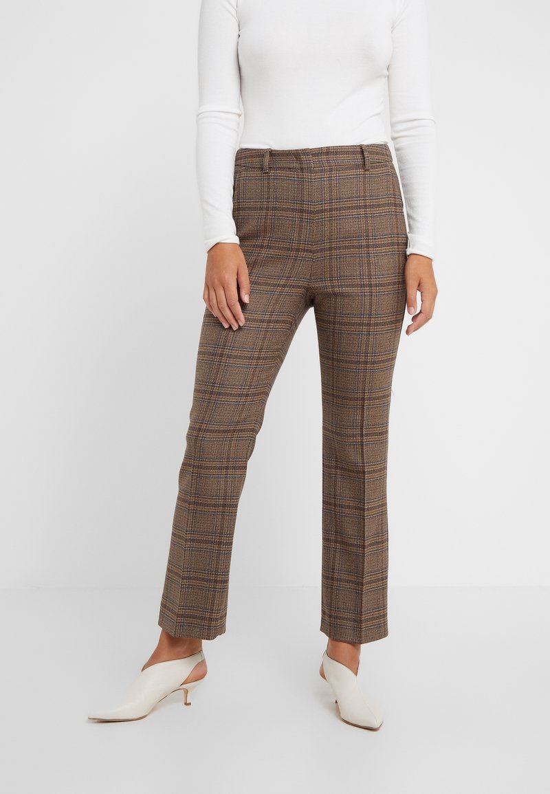 Femme portant un pantalon à jambes droites à carreaux marron, un haut blanc à manches longues et des chaussures slingback blanches à petit talon, debout devant un fond blanc uni.