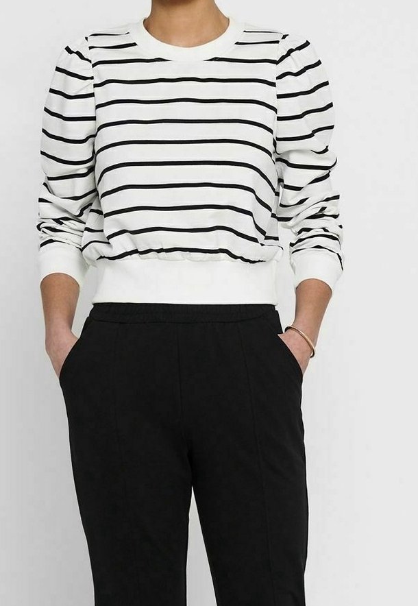 Personne portant une chemise blanche à manches longues avec des rayures horizontales noires et un pantalon noir taille haute, les mains dans les poches.