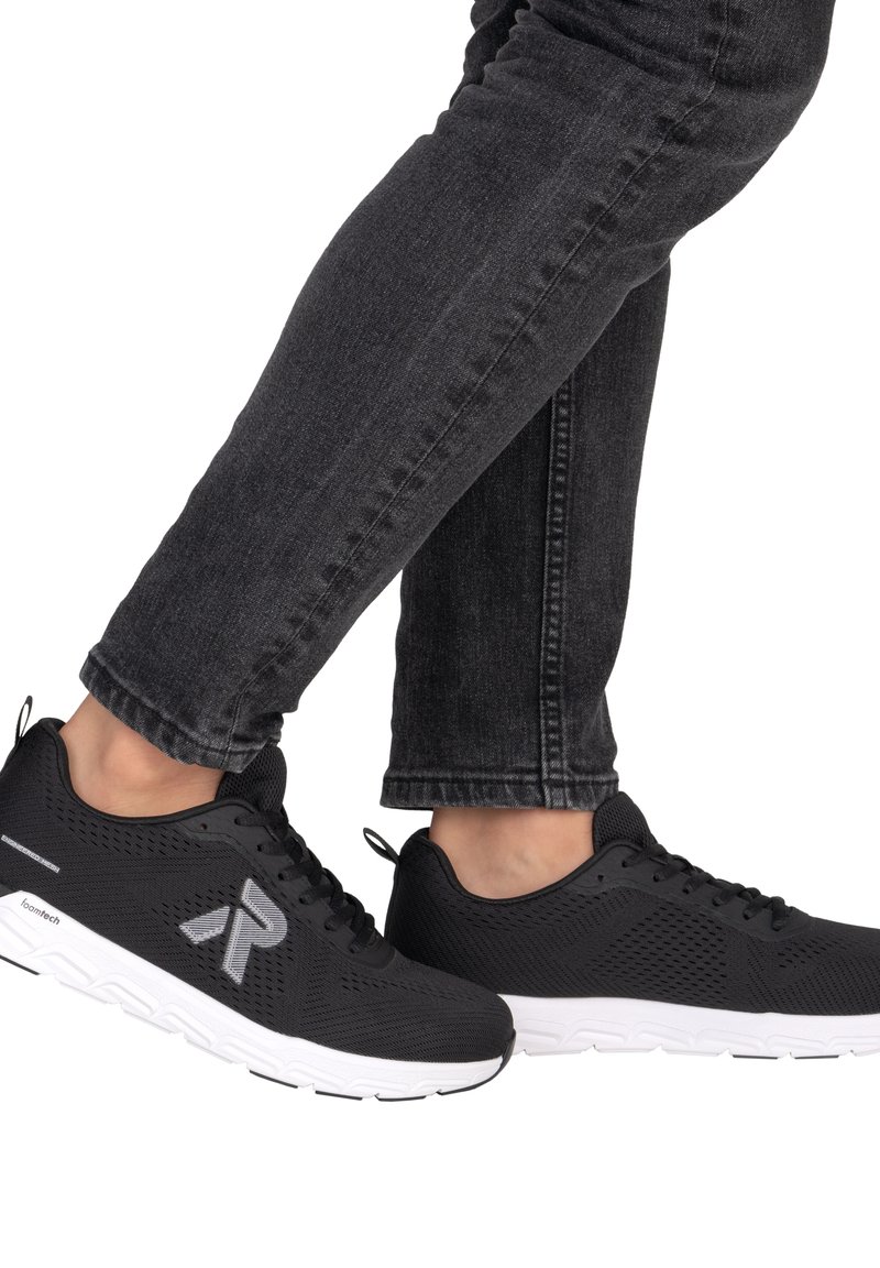 Person trägt schwarze atmungsaktive Sportschuhe mit weißen Sohlen und schwarze Jeans, zeigt eine Seitenansicht beider Füße in Gehposition.